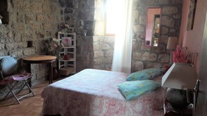 A bedroom with a stone wall, a bed with floral bedding, a small round table, a chair, and a shelf with flowers.
