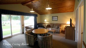 A chalet living room with a wooden ceiling, a round table, two armchairs, a sofa, a lamp, and a view of the outdoors. - The Glen House Resort (Leeds and the Thousand Islands)