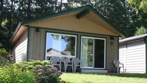 A small, single-story building with a green roof and beige siding. It features a large sliding glass door and a small porch area with white plastic chairs. - The Glen House Resort (Leeds and the Thousand Islands)