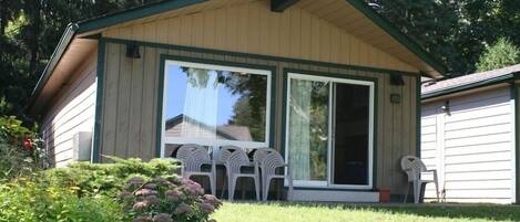 A small, single-story building with a green roof and beige siding. It features a large sliding glass door and a small porch area with white plastic chairs.