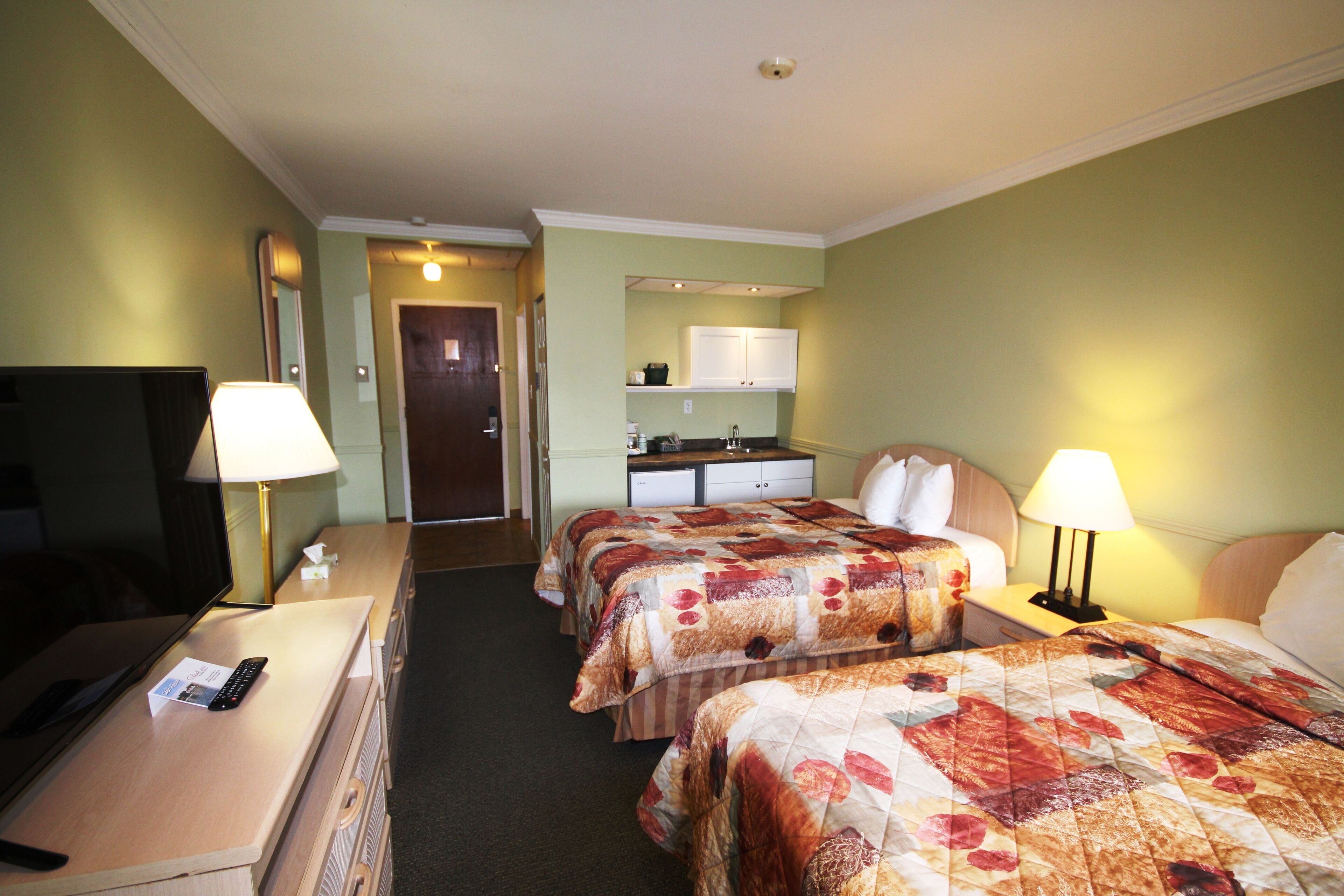 A hotel room with two beds, a desk, a TV, and a kitchenette.