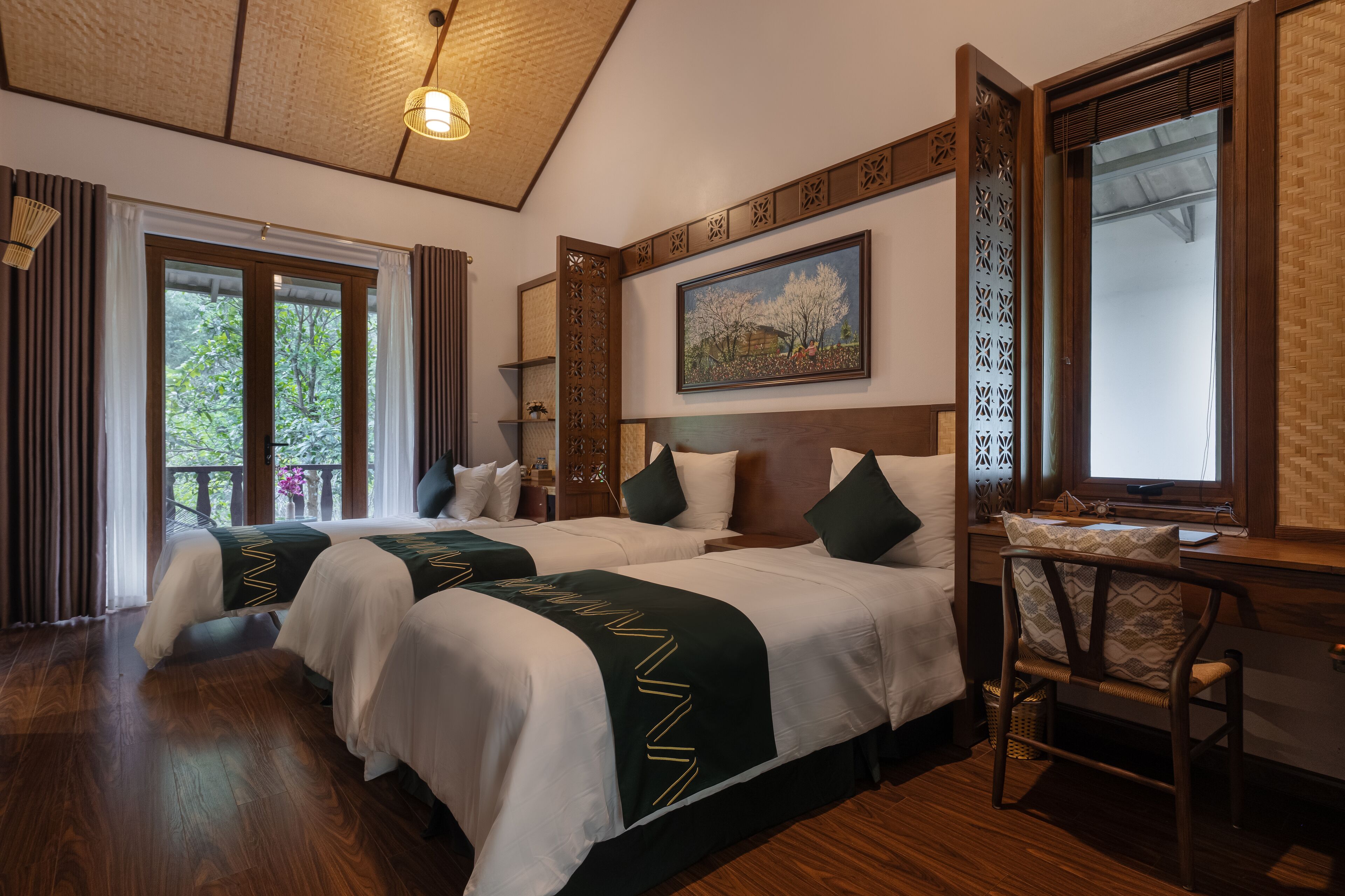 Camera d'albergo con due letti, una scrivania e una sedia. Ampia finestra con tende, un quadro appeso al muro e una ventola a soffitto.