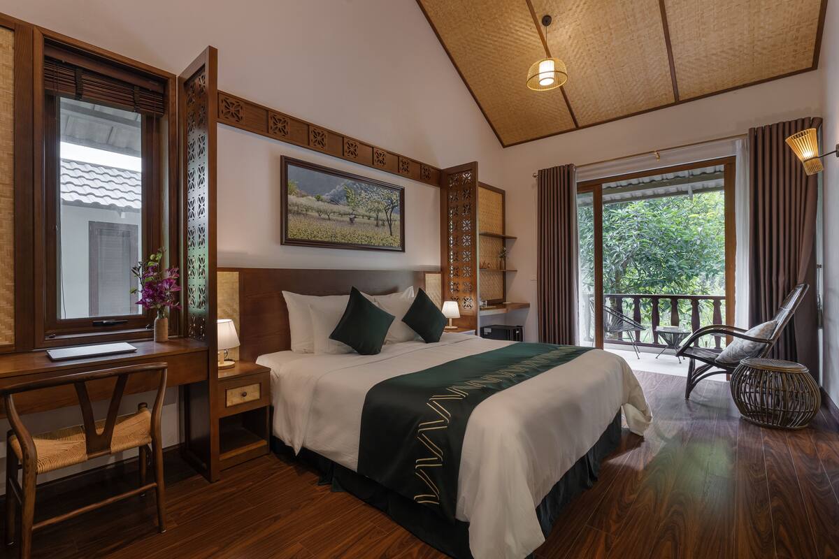 A hotel room with a large bed, a desk, a chair, and a balcony with a view of greenery.