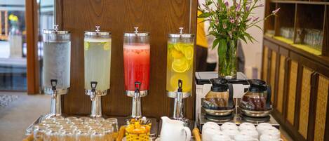 Una estación de bebidas con cuatro dispensadores, una bandeja de vasos, una jarra y un florero con flores.