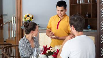 Un comedor con una mesa de madera dispuesta para tres personas, un camarero con camisa amarilla y un arreglo floral sobre la mesa.
