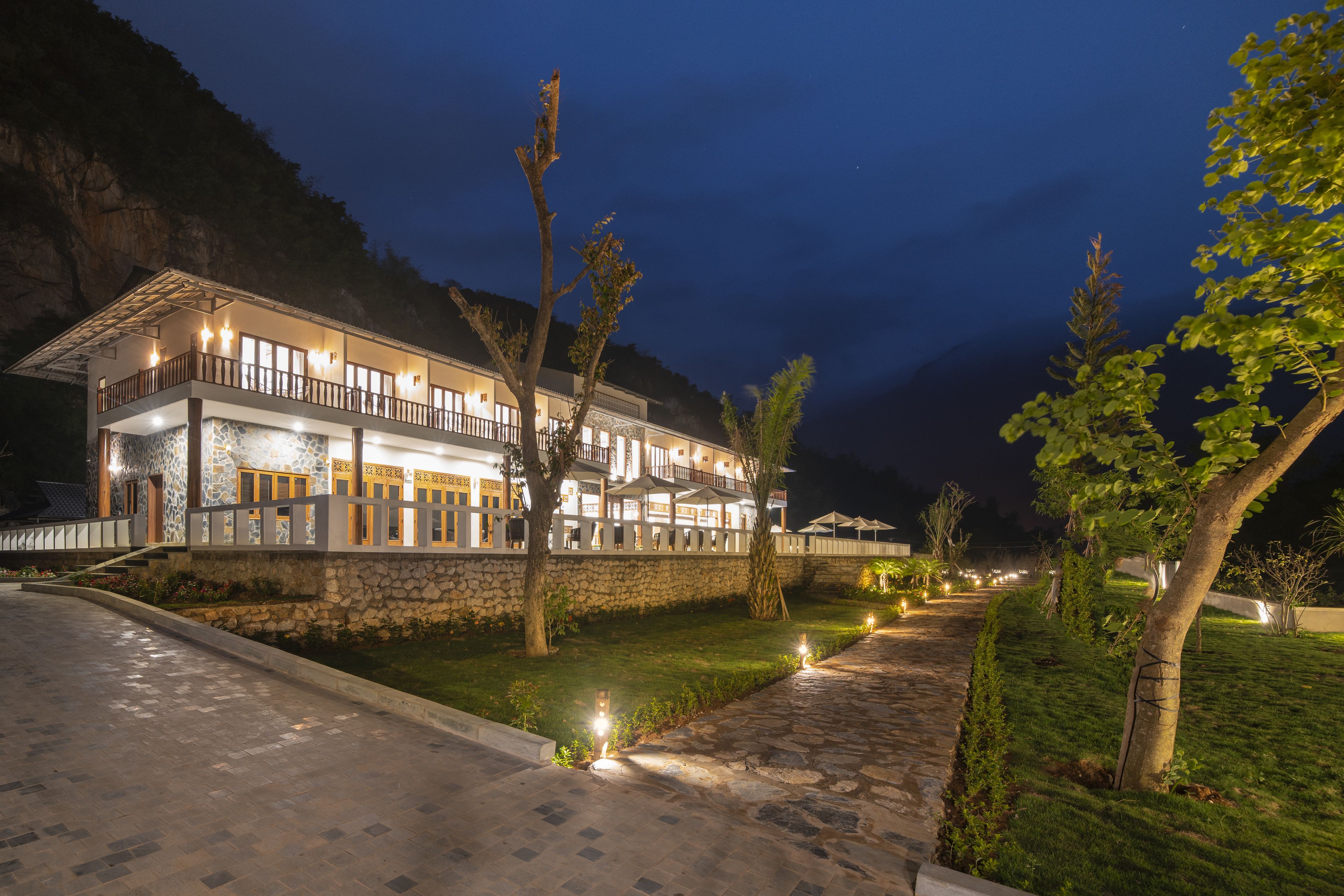 Un hotel con fachada de piedra, varios balcones y un espacio exterior bien iluminado con un camino y árboles.