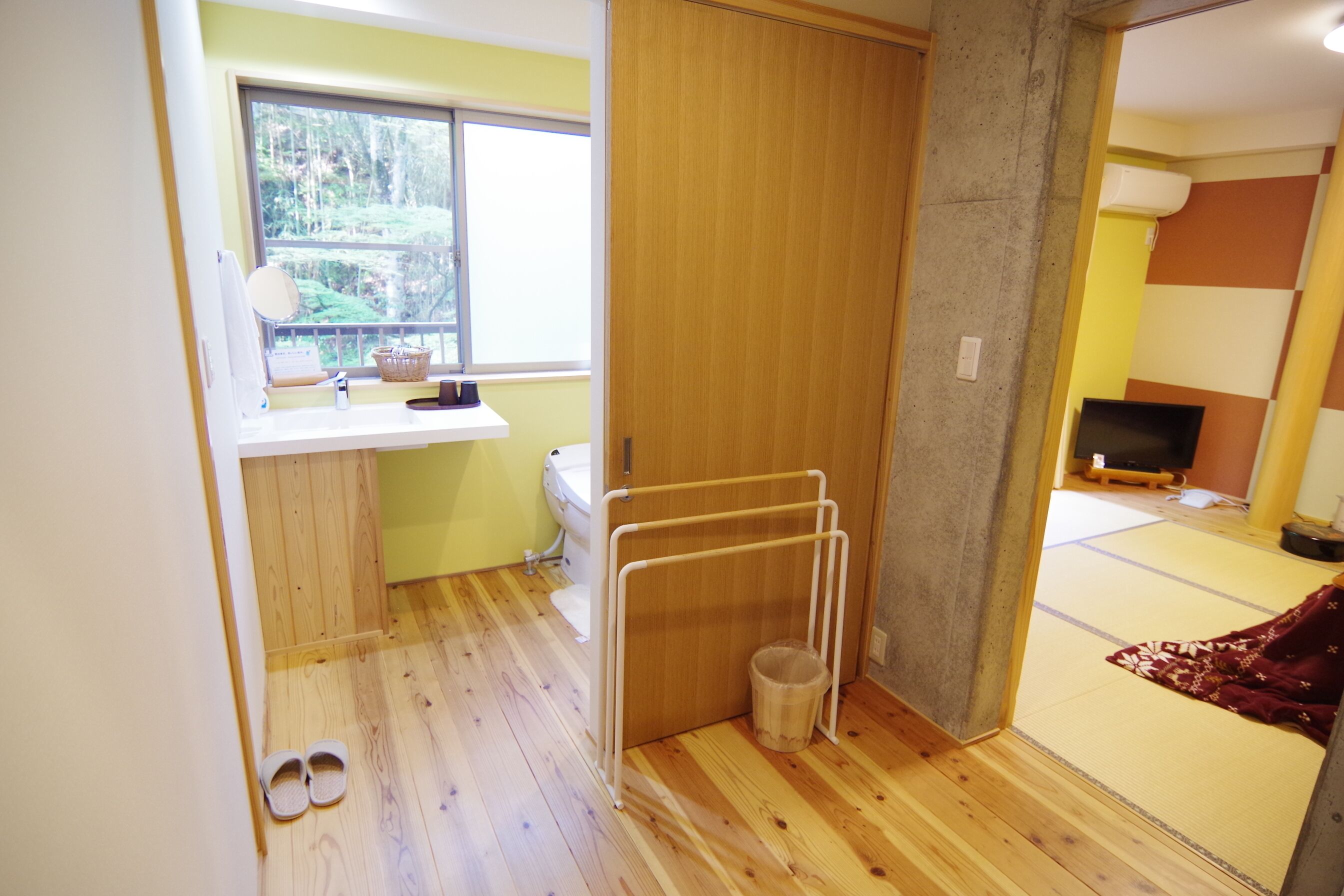 A room with a wooden floor, a window with a view of trees, a toilet, a small sink, a TV, and a tatami mat area.
