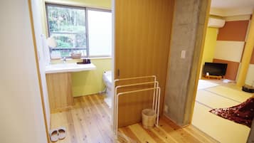 A room with a wooden floor, a window with a view of trees, a toilet, a small sink, a TV, and a tatami mat area.