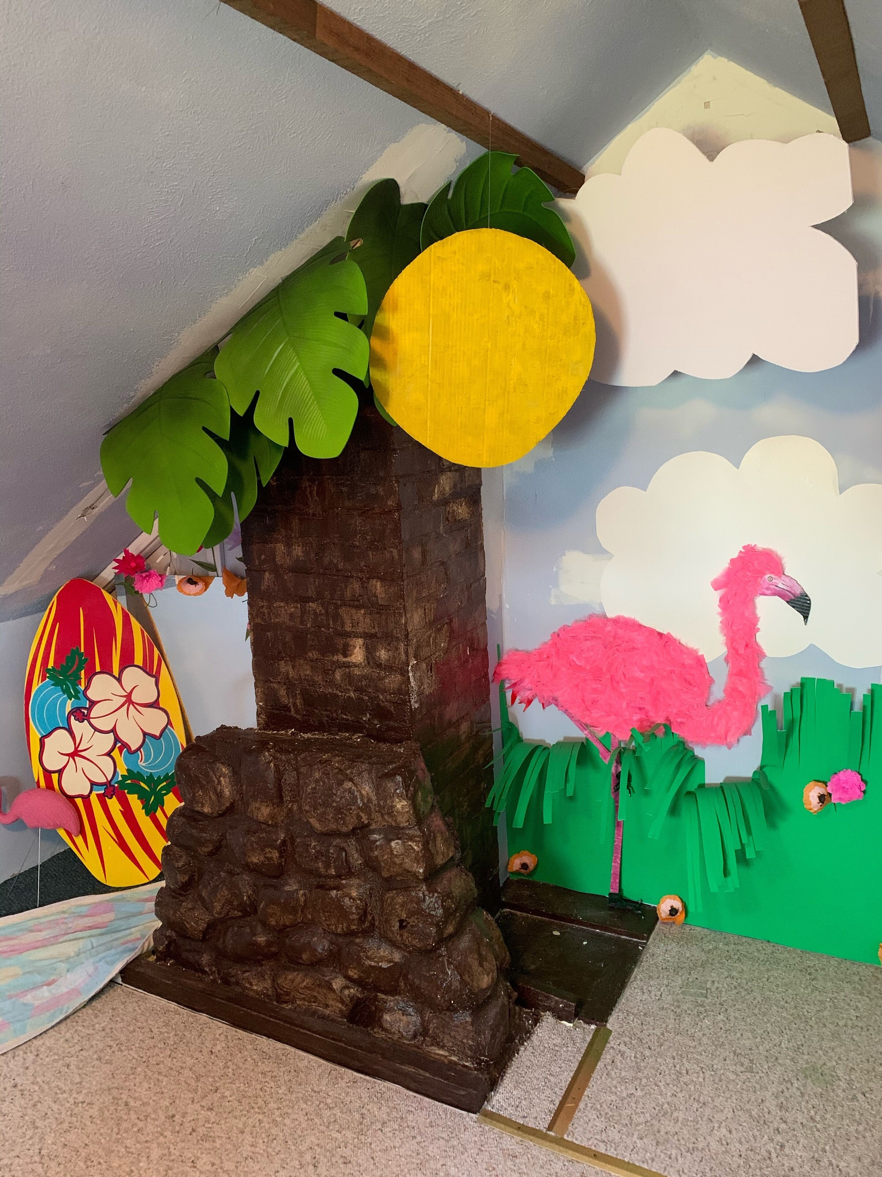 Une pièce avec un plafond en bois, un petit arbre avec une feuille jaune, un flamant rose et une planche de surf décorée de fleurs.