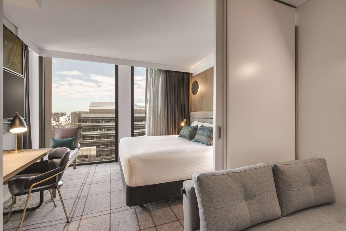 Een hotelkamer met een groot bed, een bureau, een stoel en een bank. Er is een groot raam met uitzicht op gebouwen.