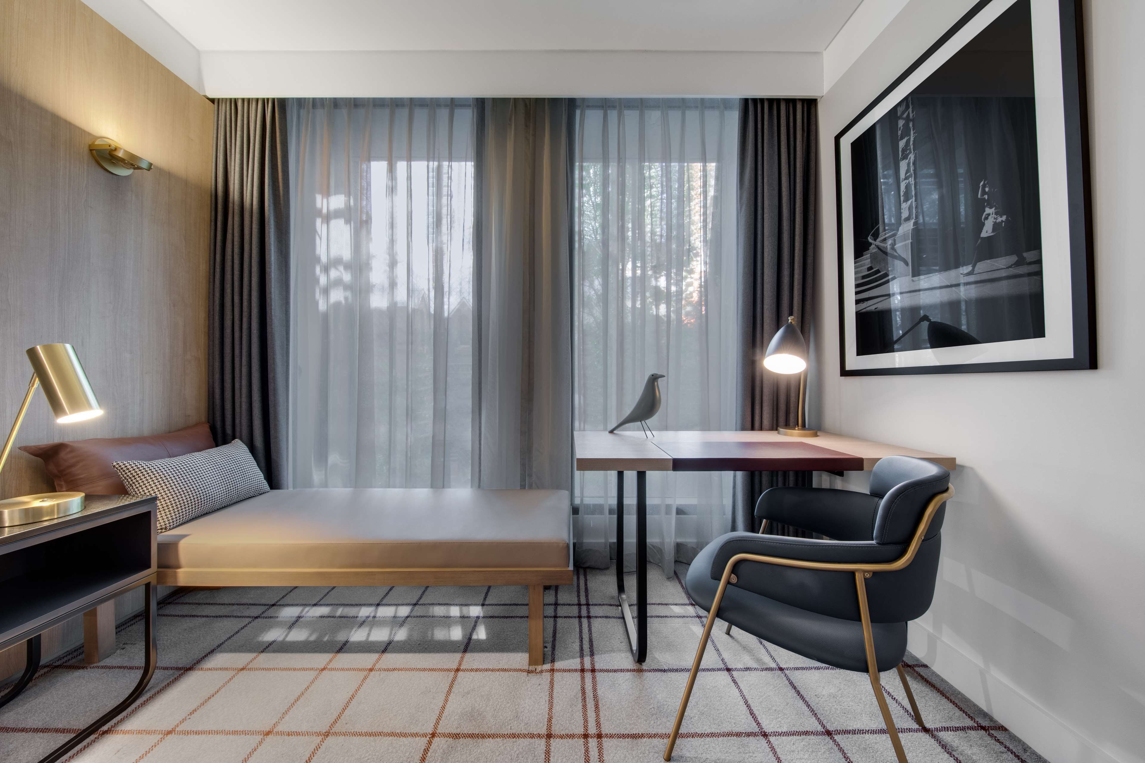 A modern hotel room with a bed, a desk, a chair, a lamp, and a framed picture on the wall.