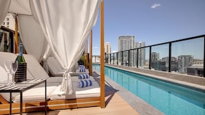 Un espace piscine aménagé sur le toit, avec des chaises longues, une table avec des verres et une bouteille, et une vue sur la ville.
