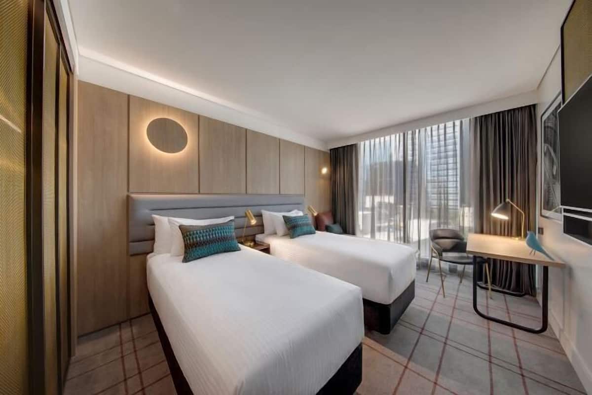 A hotel room with two beds, a desk, a chair, a TV, and a large window with curtains.