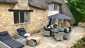 Un bâtiment en pierre avec un toit de chaume, une terrasse dotée de meubles en osier et un petit jardin.