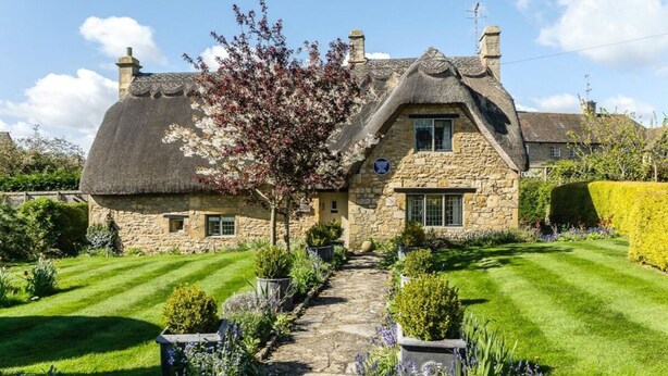 Une maison en pierre avec un toit de chaume, un chemin de pierre et des jardins bien entretenus.