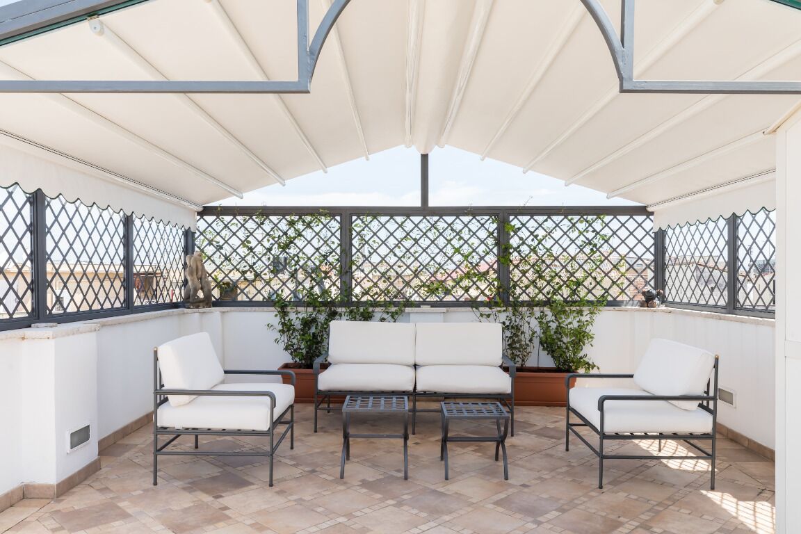 Un espace extérieur aménagé, abrité et doté de chaises et de tables blanches avec des coussins, entouré d’une clôture en métal et agrémenté de plantes en pot.