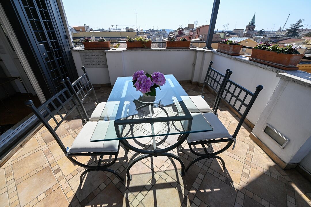 A glass-top table with four chairs on a tiled balcony, a vase with purple flowers, and potted plants on the railing.