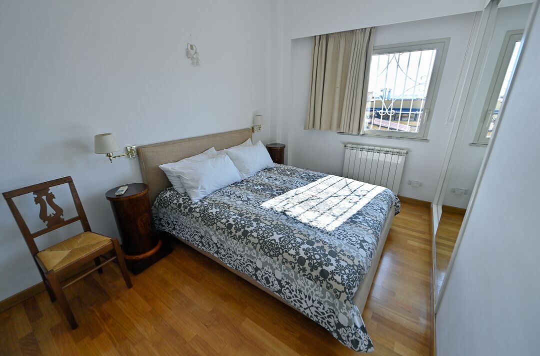 A bedroom with a bed, a chair, a nightstand, a window with curtains, and a radiator.