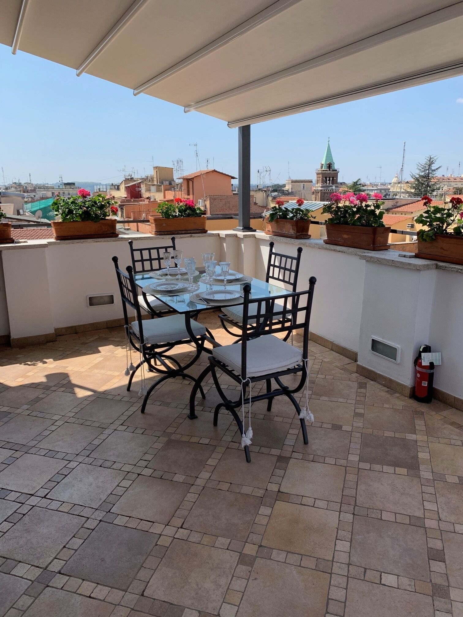 Una terraza en la azotea con una mesa preparada para comer, rodeada de plantas en macetas y con vistas a un paisaje urbano.
