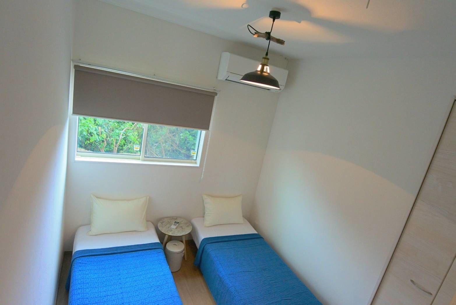 A small, single-room accommodation with two beds, a small round table, a window with a view of greenery, and a ceiling-mounted light fixture.