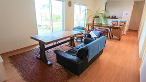 Un salón espacioso con una mesa de madera, un sofá de cuero negro y una alfombra roja. Se ven dos ventanas con vistas a zonas verdes.