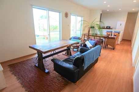 A spacious living room with a wooden table, a black leather sofa, and a red rug. There are two windows with a view of greenery outside.
