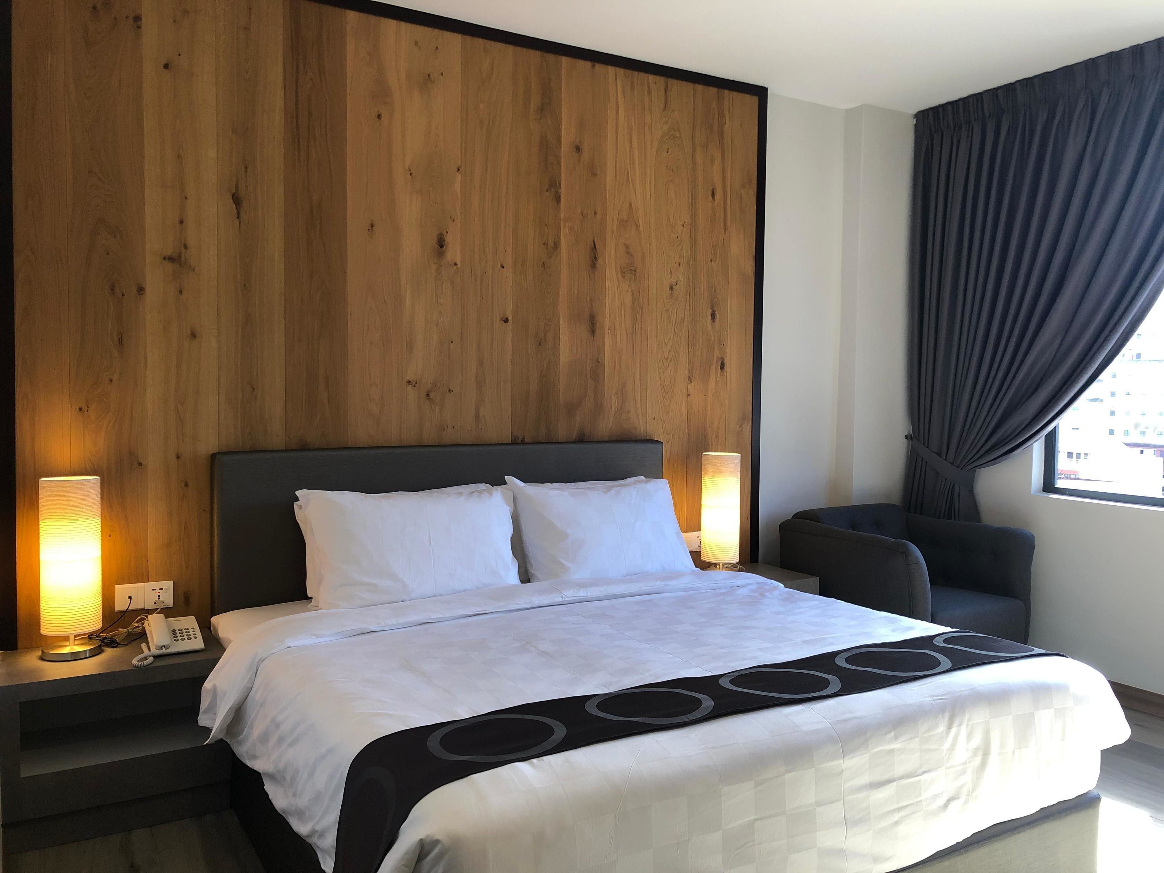 A modern hotel room with a wooden panel headboard, a large bed with white bedding, two bedside lamps, a bedside phone, and a view of the city through a window with dark curtains.