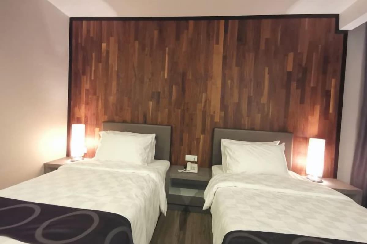 A hotel room with two beds, wooden headboard, bedside lamps, and a small table with a phone.