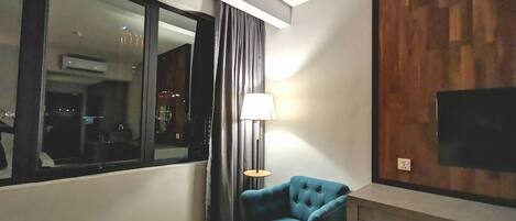A modern living room with a large window, a blue armchair, a lamp, and a flat-screen TV mounted on the wall.
