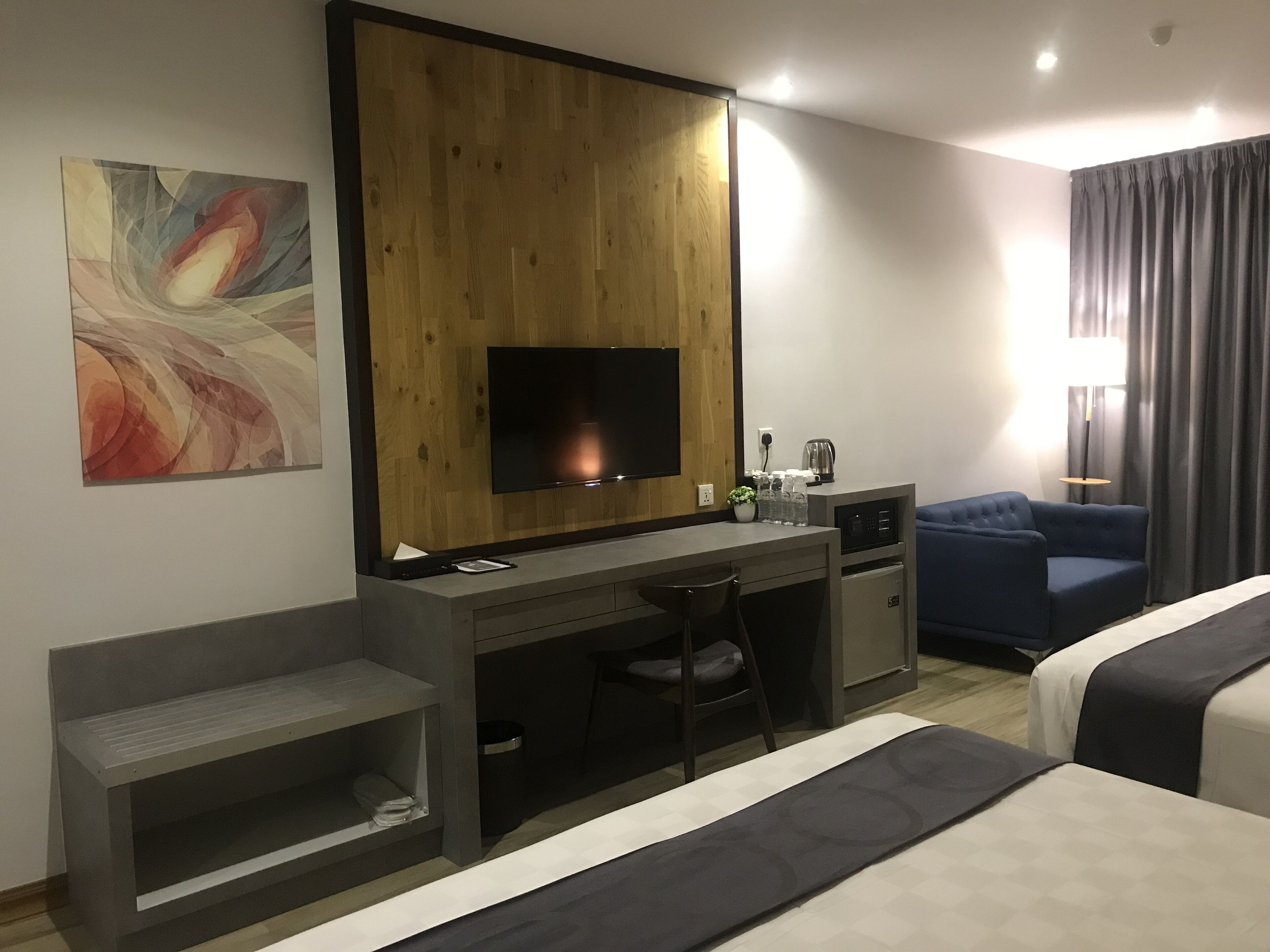 A modern hotel room with a wooden accent wall, a flat-screen TV, a desk with a chair, a sofa, and a bed.