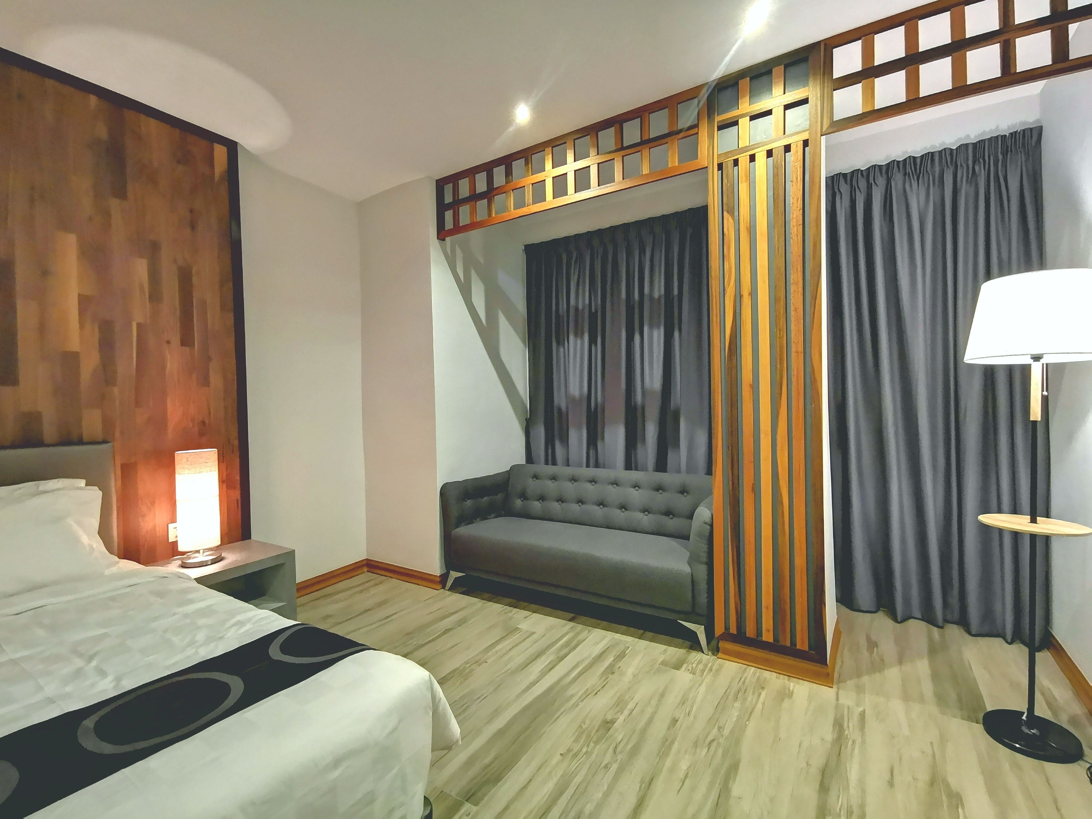 A hotel room with a bed, a nightstand, a lamp, a sofa, and a large window with curtains.