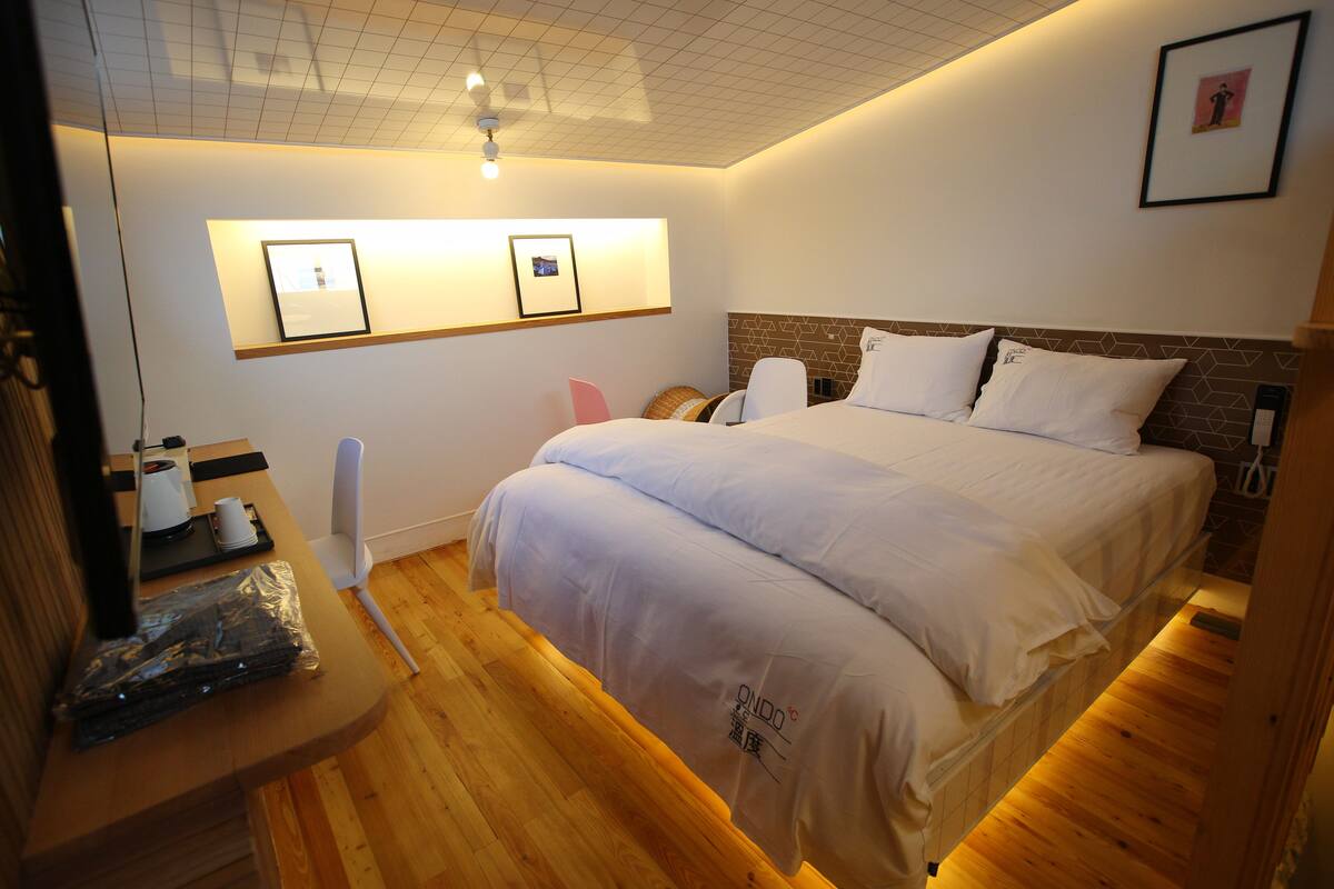 Una habitación de hotel ordenada con una cama grande, suelo de madera y un estante en la pared con un cuadro enmarcado y una pequeña mesa con una taza y una caja de pañuelos.