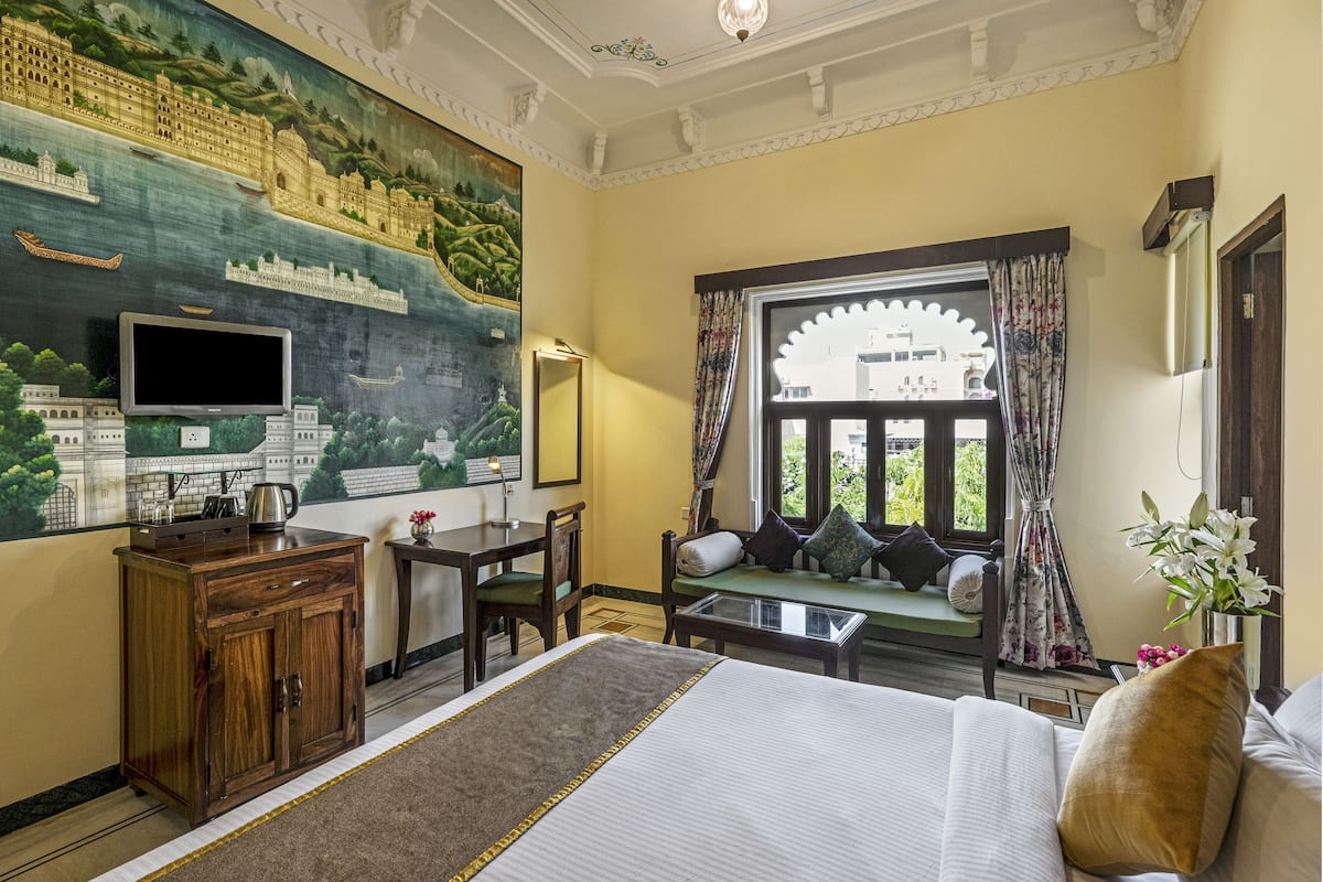 Une chambre d’hôtel avec une grande fresque murale, un lit, un bureau avec une chaise, une petite table et une fenêtre avec des rideaux.