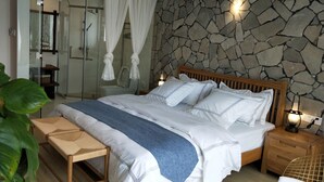 A bedroom with a stone wall, a wooden bed, a bench, and a bedside table with a lamp. - 25 Degree Sea Boutique Homestay (Zhoushan)