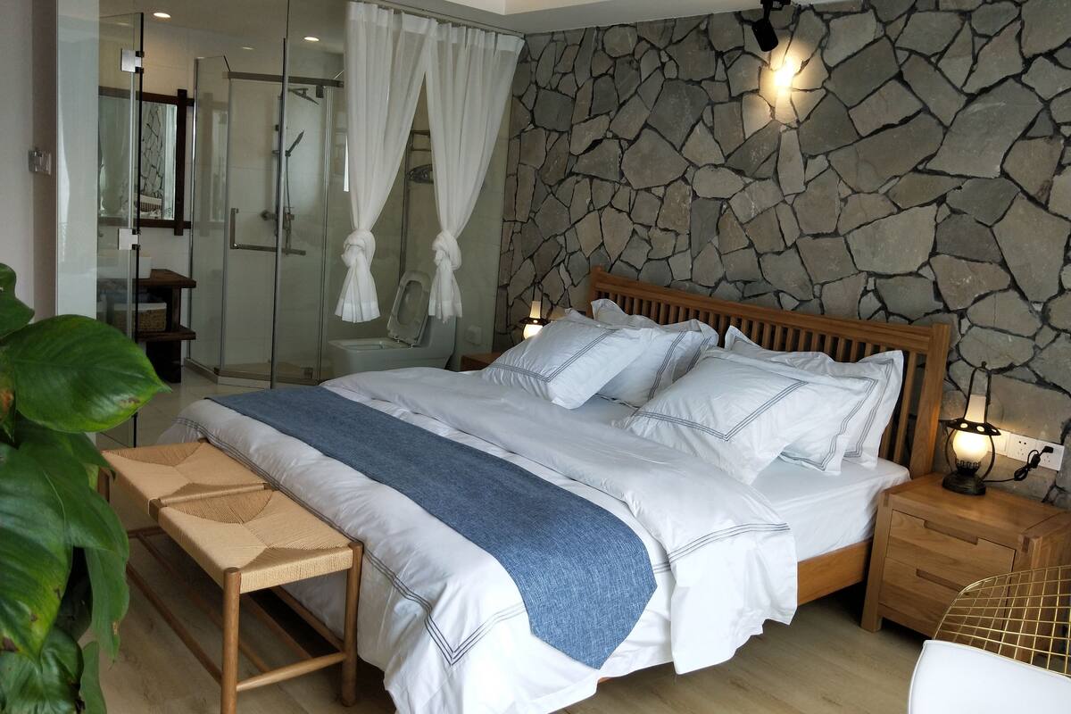 Un dormitorio con pared de piedra, cama de madera, un banco y una mesita de noche con lámpara.