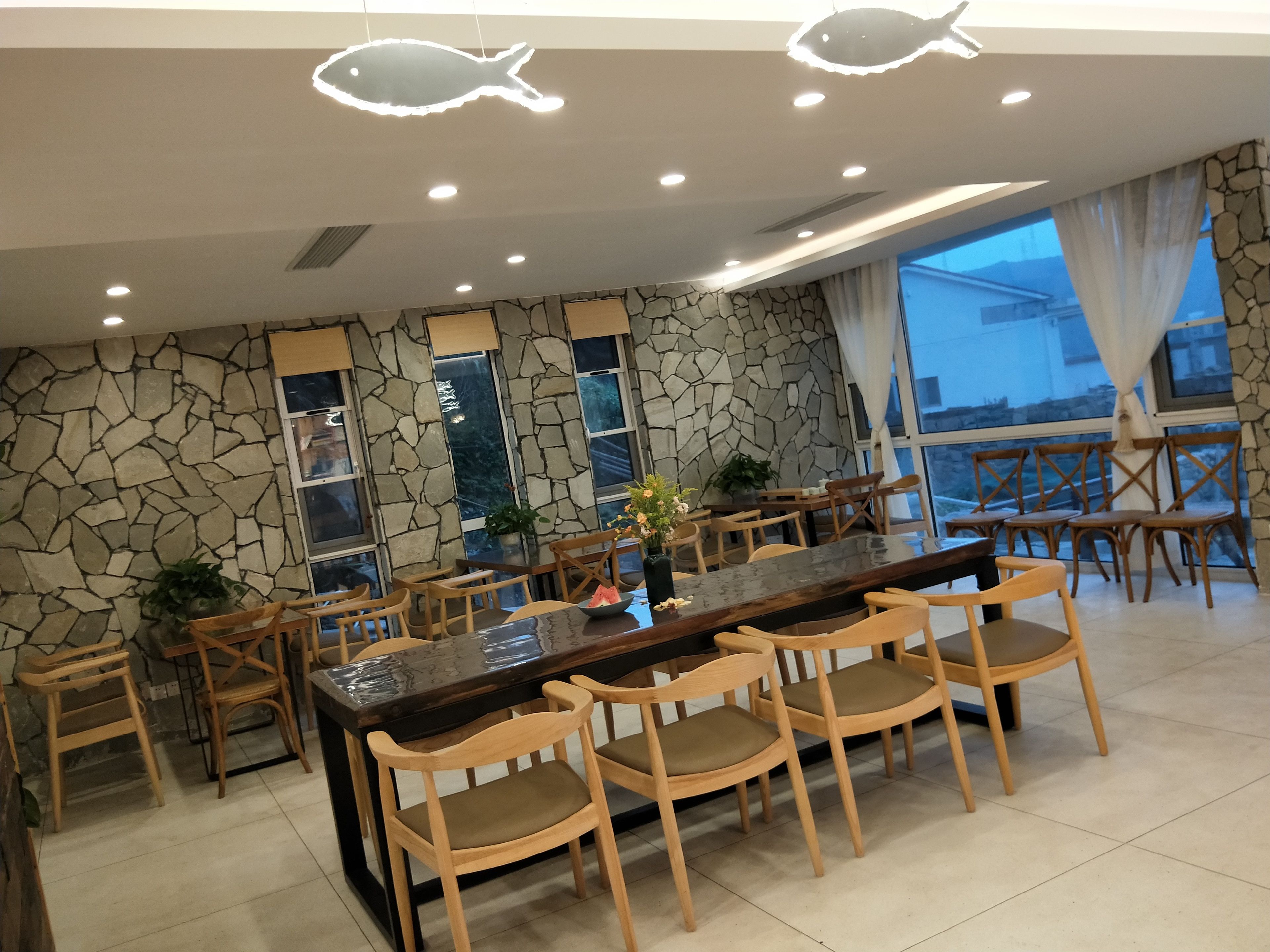A restaurant with a stone wall, wooden chairs, a long table, and a fish-shaped light fixture.