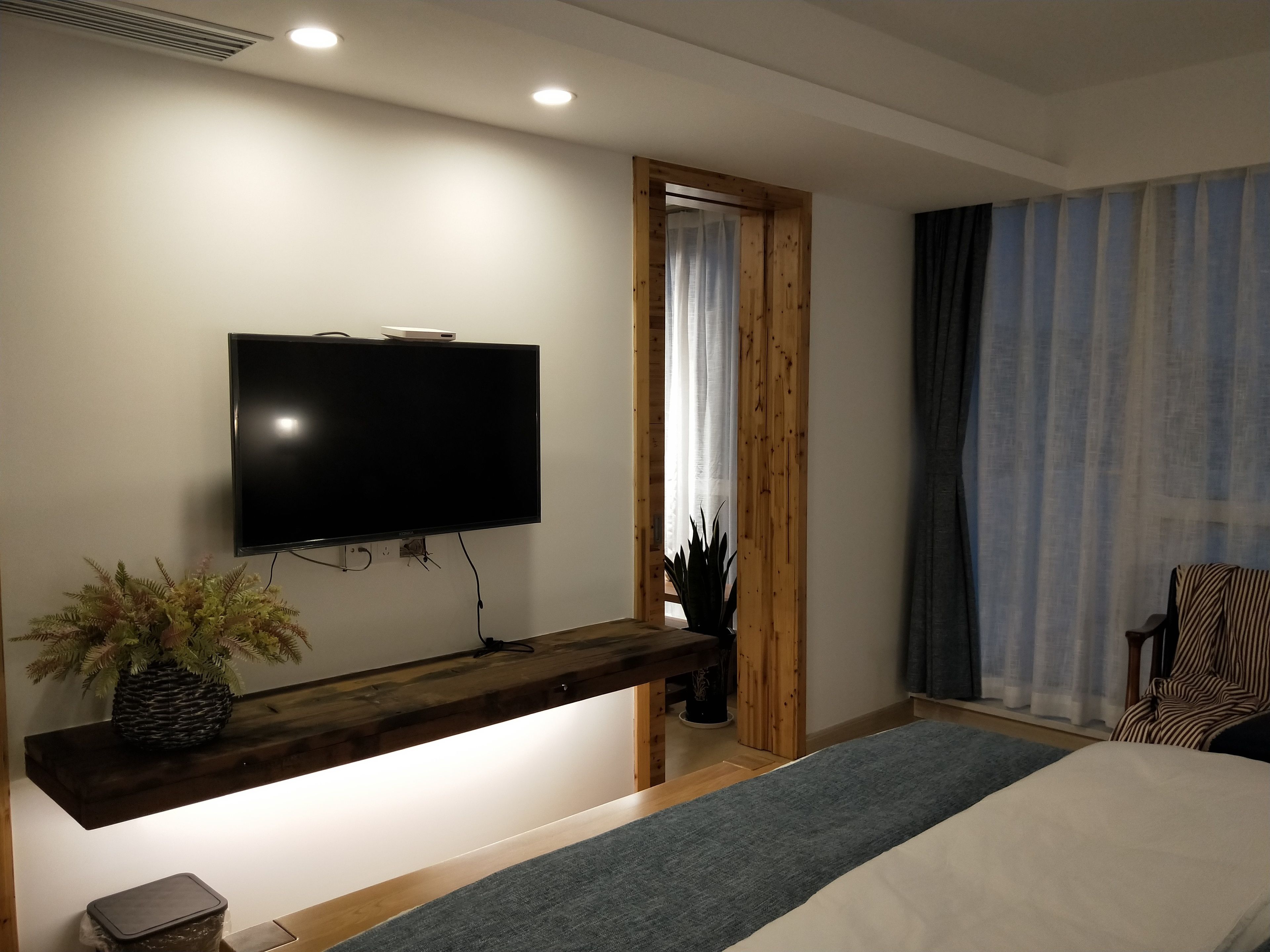 A modern hotel room with a flat-screen TV mounted on a wooden console, a bed with a grey runner, a potted plant, and a window with curtains.