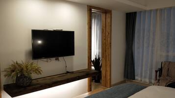 A modern hotel room with a flat-screen TV mounted on a wooden console, a bed with a grey runner, a potted plant, and a window with curtains.