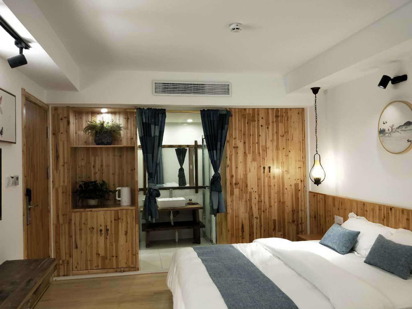 A bedroom with a wooden wardrobe, a bed with pillows, a bedside table, a hanging light fixture, and a wall-mounted mirror.