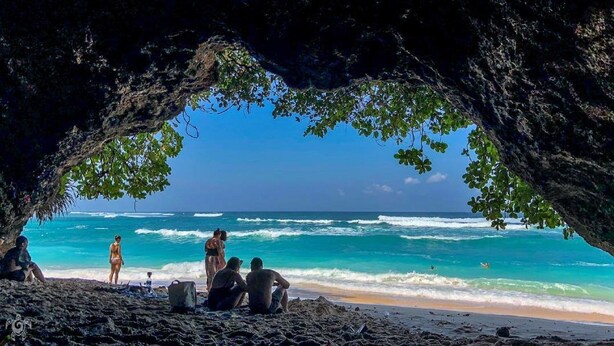 A beach scene with a cave entrance, people sitting and standing on the sand, and the ocean in the background. - Bali Bobo Hostel (Jimbaran)