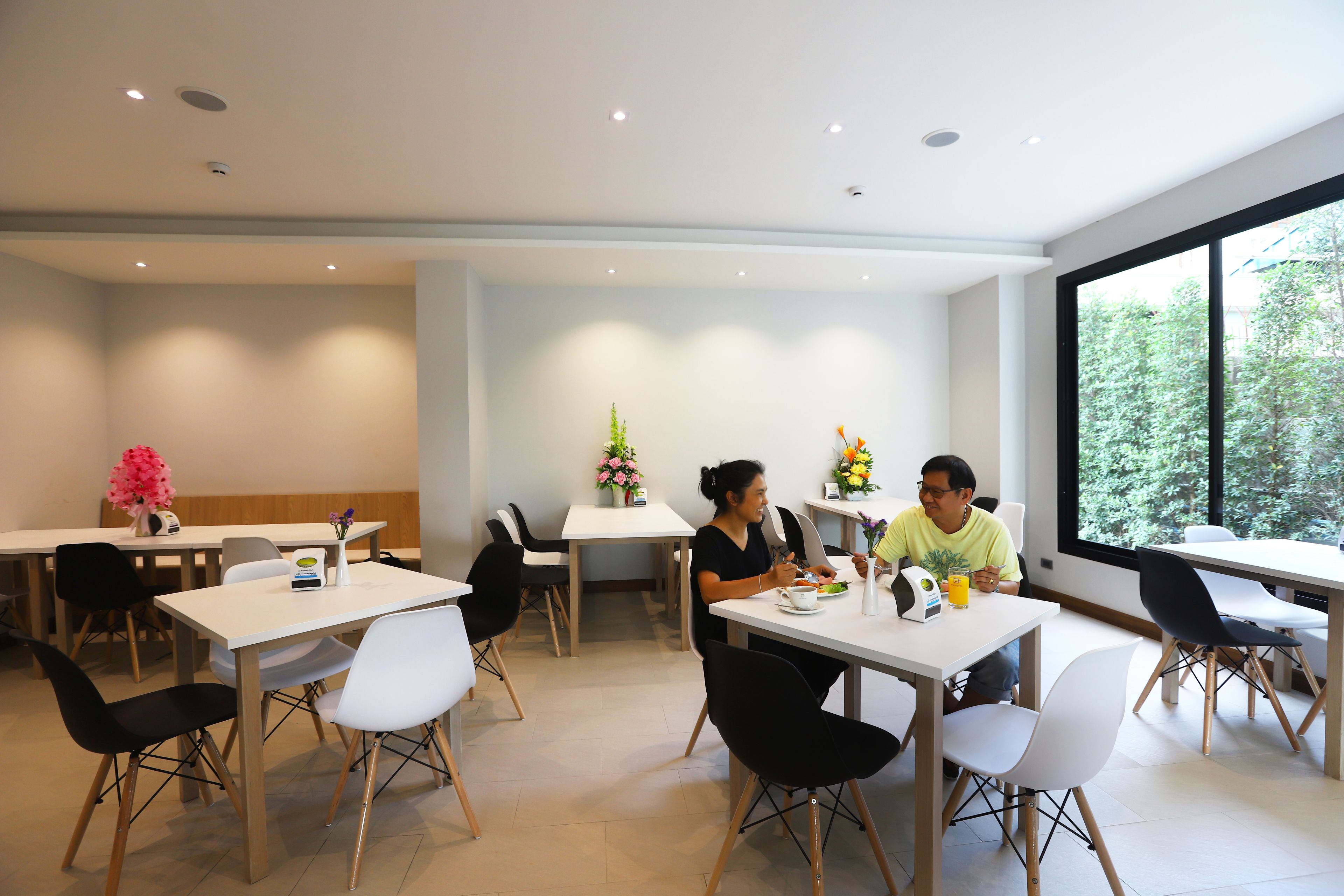 A modern dining area with white tables and chairs, a large window with a view of trees, and a ceiling with recessed lighting.