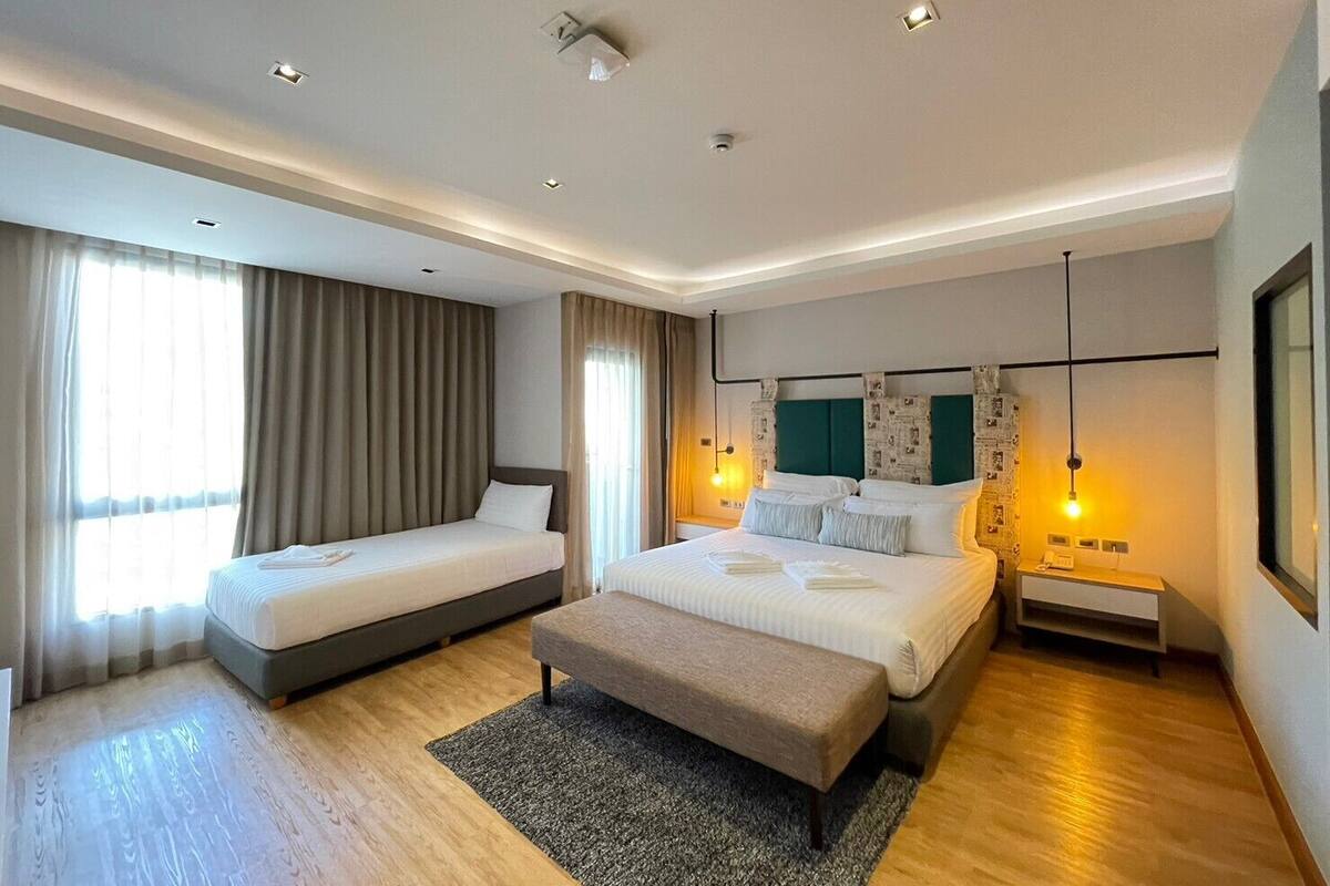 A modern hotel room with two single beds, a larger bed, a bench, a desk, and a nightstand.
