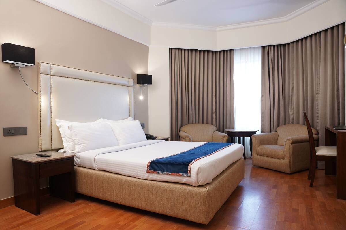 Una habitación de hotel con una cama grande, una mesita de noche con lámpara, una silla, una pequeña mesa y un televisor montado en la pared.