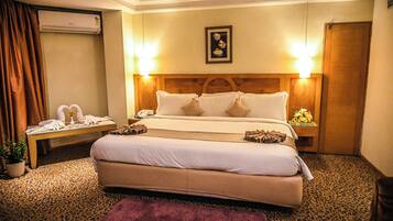 A hotel room with a large bed, a bedside table with a lamp, a potted plant, and a wall-mounted air conditioner.