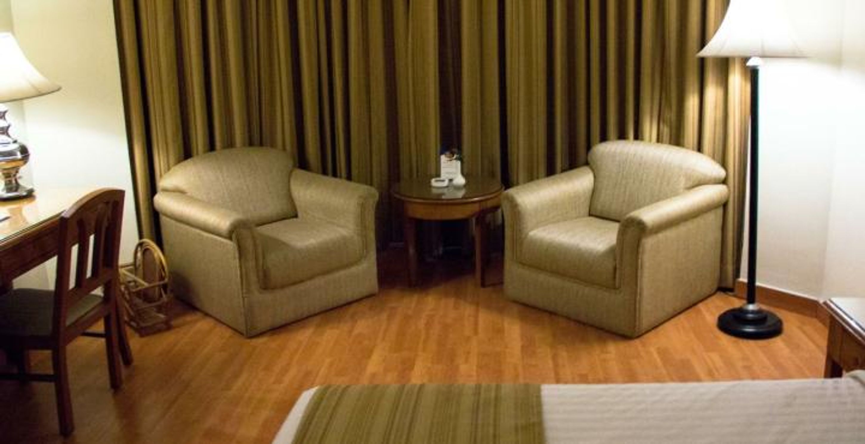 A hotel room with two armchairs, a small round table, a lamp, and a bed with a striped bedspread.