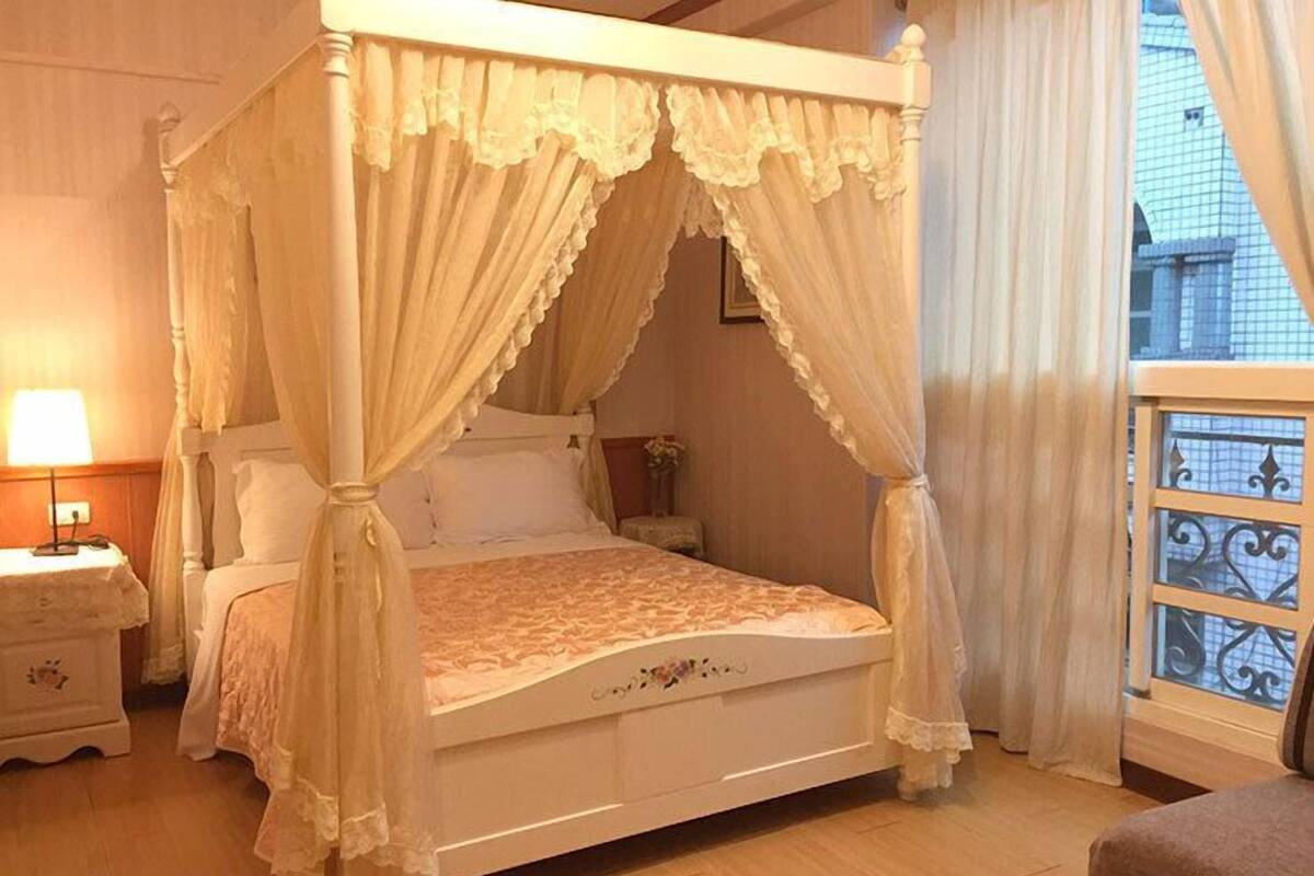 Ein Bett mit Baldachin, bedeckt mit einer Spitzenüberwurfdecke, ein Nachttisch mit Lampe und Blick auf die Stadt durch ein Fenster mit dekorativem Gitter.