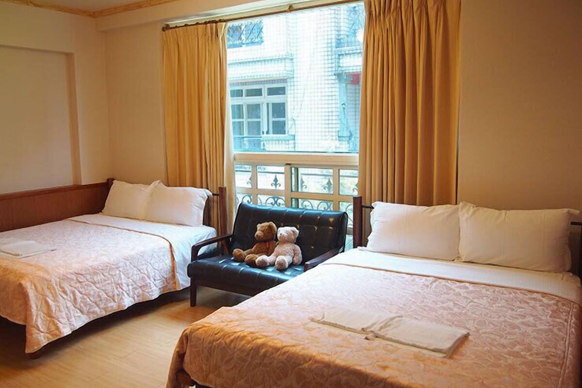 Ein Hotelzimmer mit zwei Einzelbetten, einem kleinen Sofa mit Teddybär und einem Fenster mit Vorhängen.