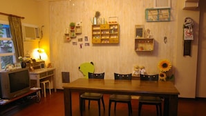 A dining area with a wooden table and chairs, a television on a cart, and wall-mounted shelves with various items. - Riverbank Cottage (Wujie)