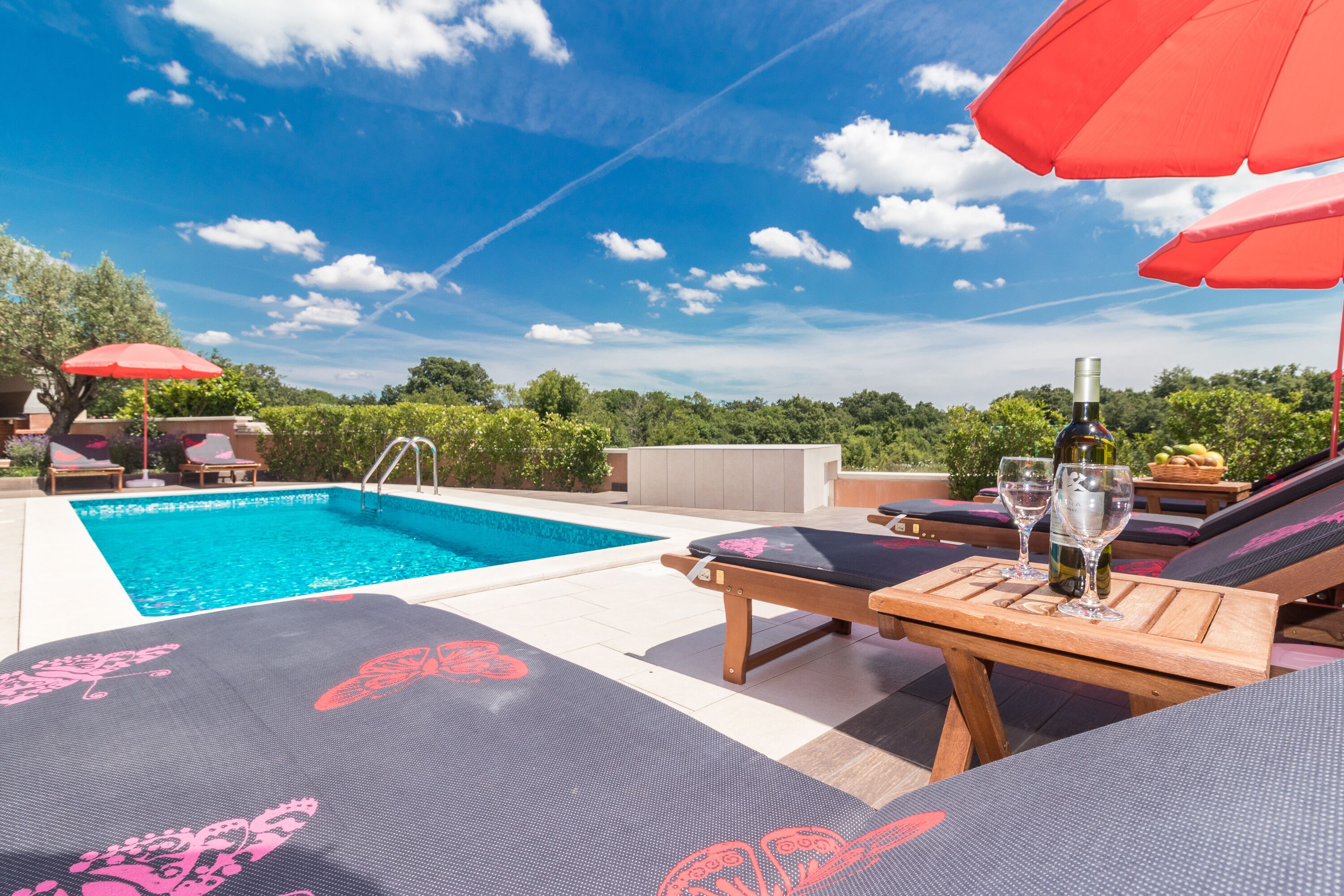 Un espace aménagé au bord de la piscine, avec des chaises longues, une table en bois et des verres à vin, le tout ombragé par un parasol rouge.