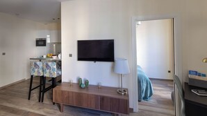 Un salon moderne avec un téléviseur à écran plat fixé au mur, une petite console en bois, un coin repas avec une nappe à motifs et un coin cuisine équipé d’un micro-ondes et d’un saladier rempli de fruits.